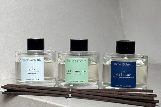 Three small clear glass perfume bottles with black caps and blue, green, and white labels are mounted on a white wall next to a bundle of dark reed diffuser sticks.