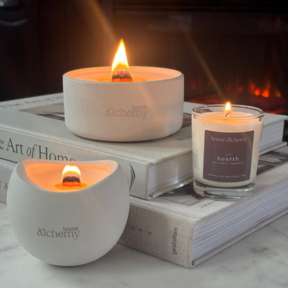 Two lit candles from the brand home Alchemy are styled on stacked books in front of a warm fireplace. One candle is in a matte white ceramic holder and the other is in a clear glass jar with a gray label reading 'hearth'.
