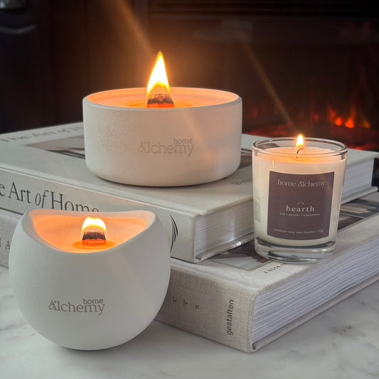 Two lit candles from the brand home Alchemy are styled on stacked books in front of a warm fireplace. One candle is in a matte white ceramic holder and the other is in a clear glass jar with a gray label reading 'hearth'.